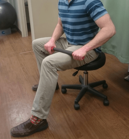 A man is seated on a rolling stool. He is rolling a rolling pin along the middle of his quadriceps (mid-thigh). 